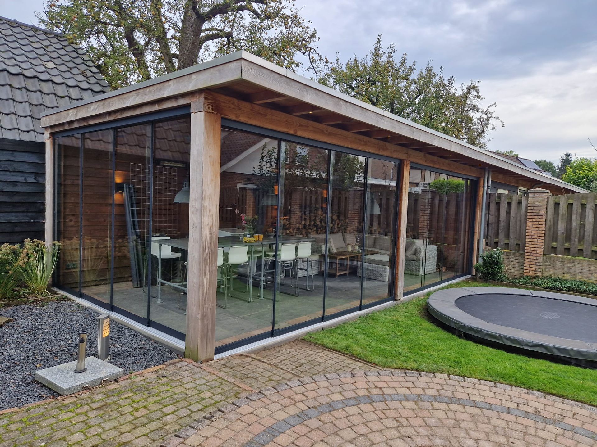 Een groot houten tuinhuisje met glazen schuifdeuren en een trampoline in de achtertuin.