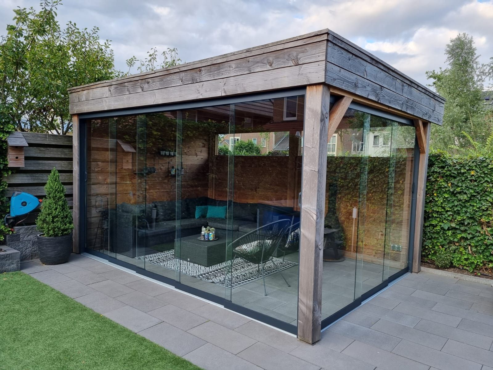 Een houten tuinhuisje met glazen schuifdeuren in een achtertuin.