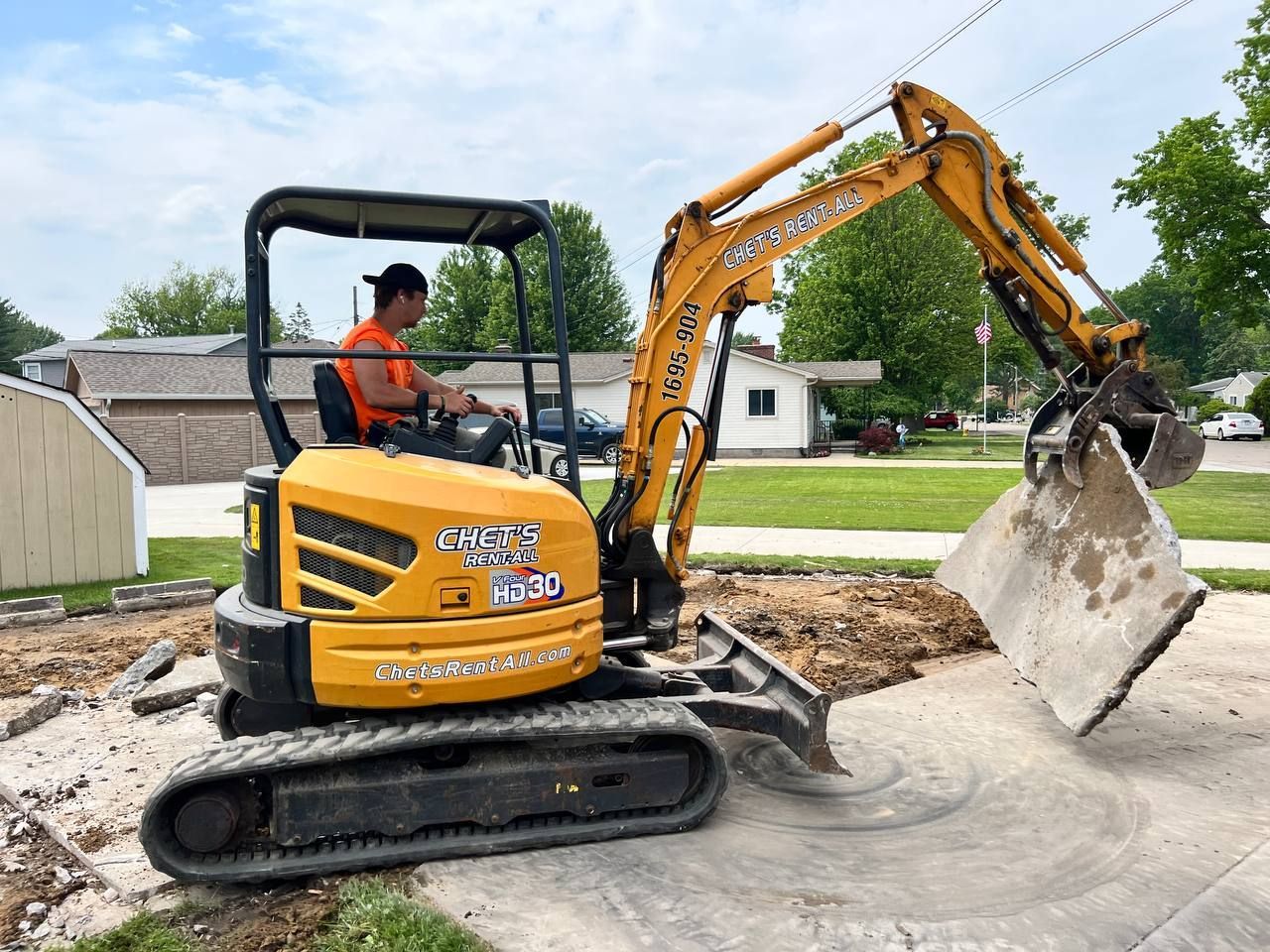 A man is driving a yellow excavator in a driveway.