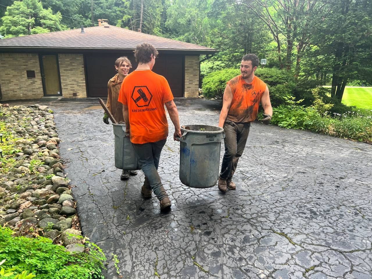 Three men are carrying buckets down a driveway in front of a house.