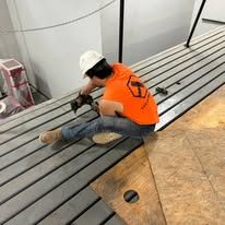 A man in an orange shirt and hard hat is kneeling on a wooden deck.
