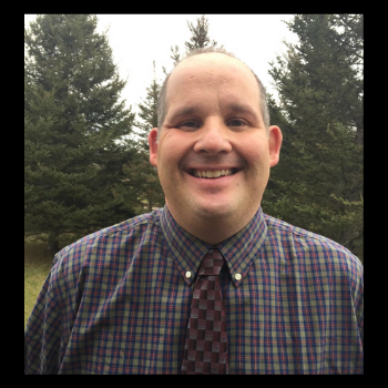 A man in a plaid shirt and tie is smiling in front of trees.