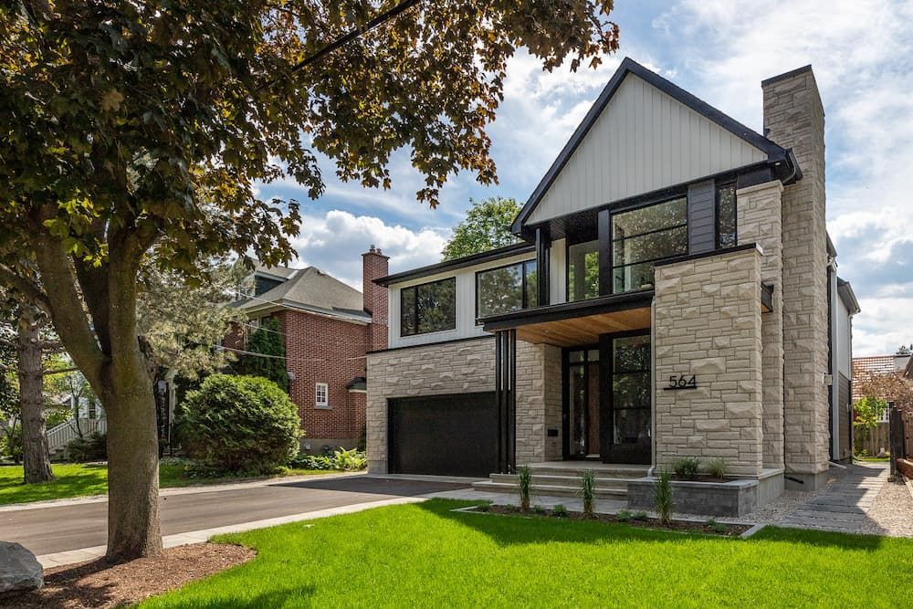 Modern two-story house with stone facade, black trim, and garage. Green lawn and mature trees.