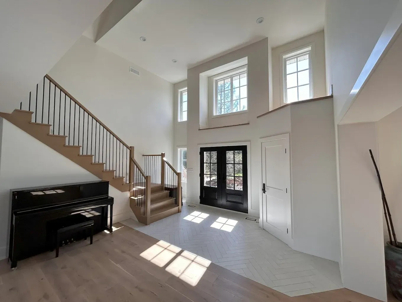 Bright entry hall with stairs, piano, and double front doors; sunlight streams in.