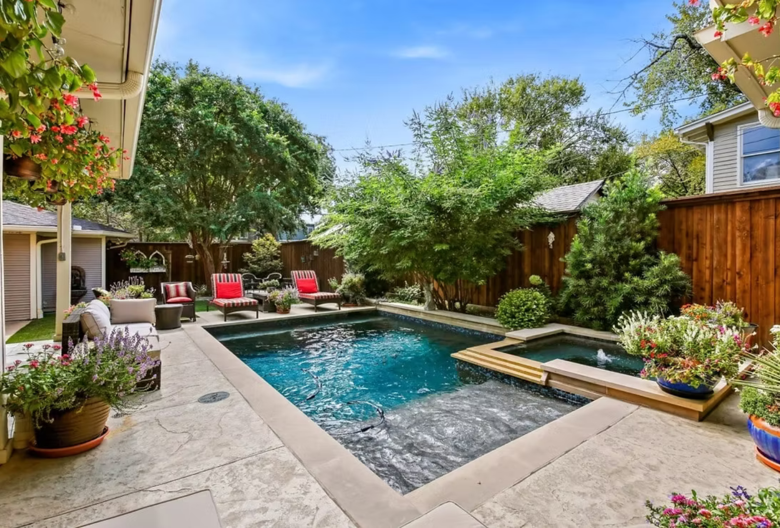 Backyard with pool and hot tub, surrounded by greenery, red chairs, and blue sky.