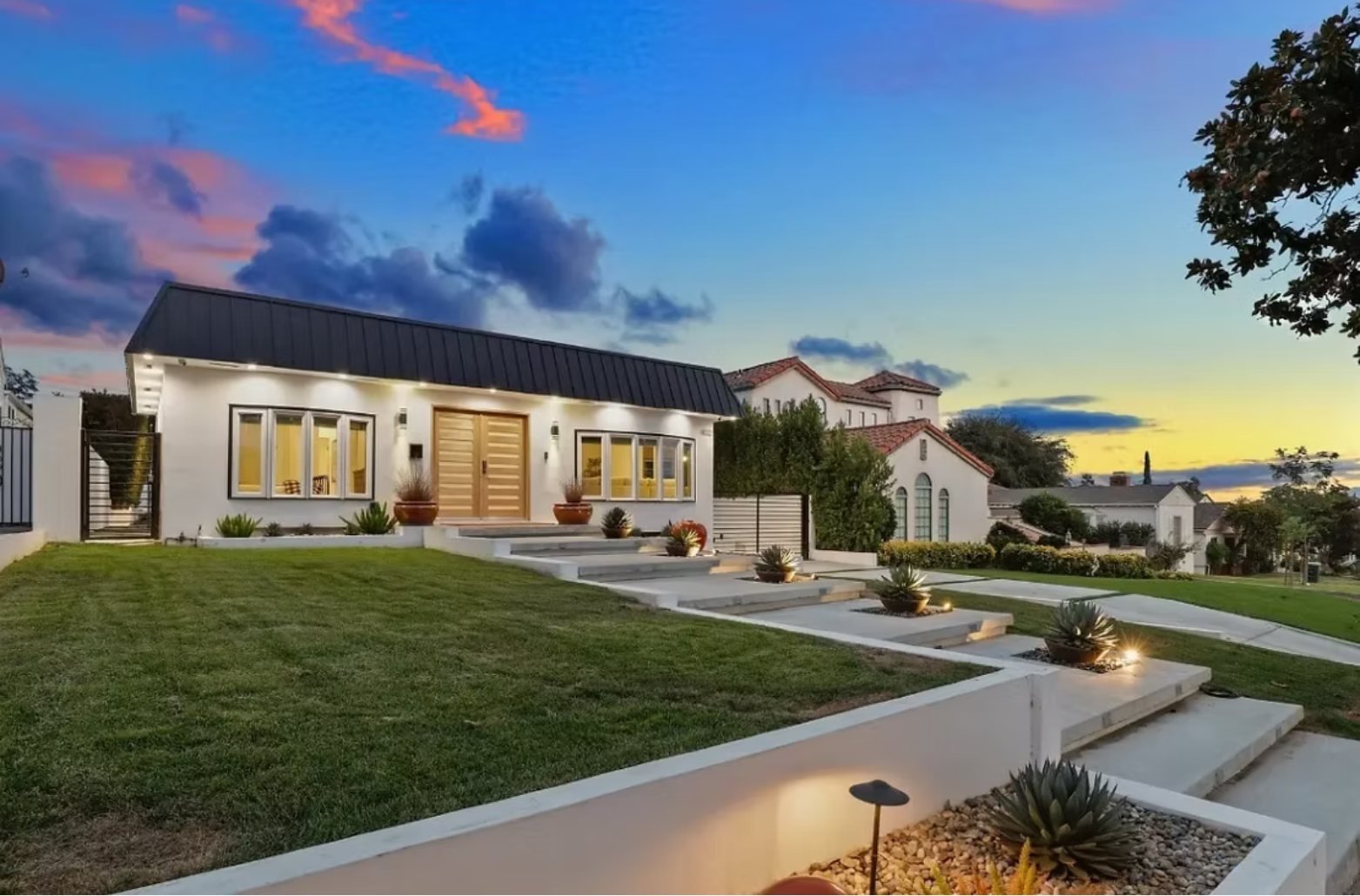 White modern house with black roof, concrete steps, and a grassy yard at dusk.