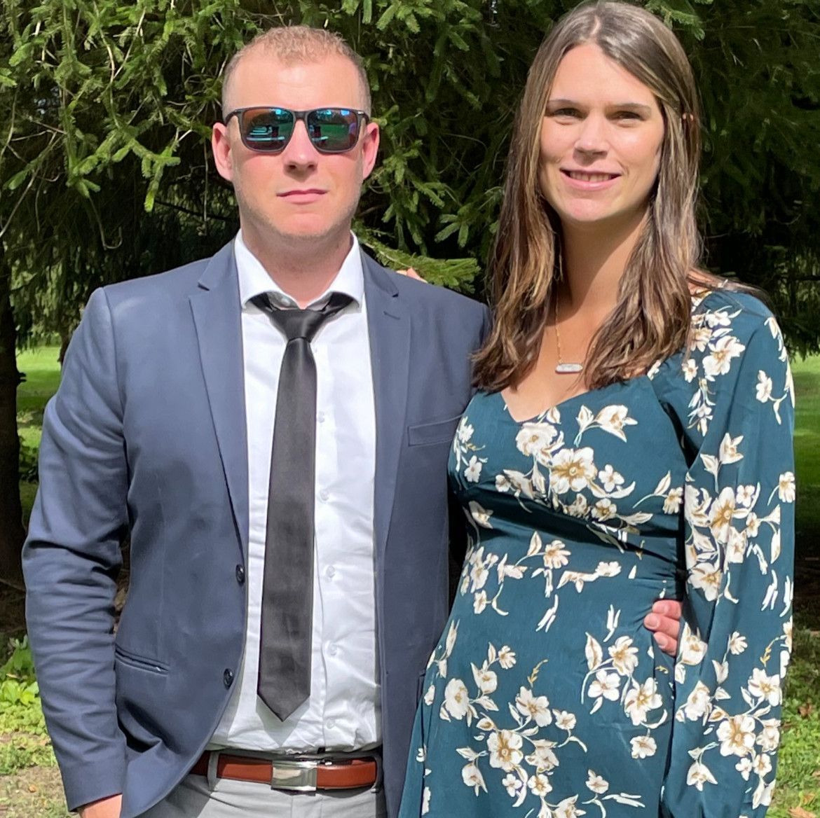 A man and woman stand together outside. The man wears sunglasses and a suit. The woman wears a floral dress.