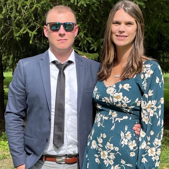 A man and woman stand together outside. The man wears sunglasses and a suit. The woman wears a floral dress.