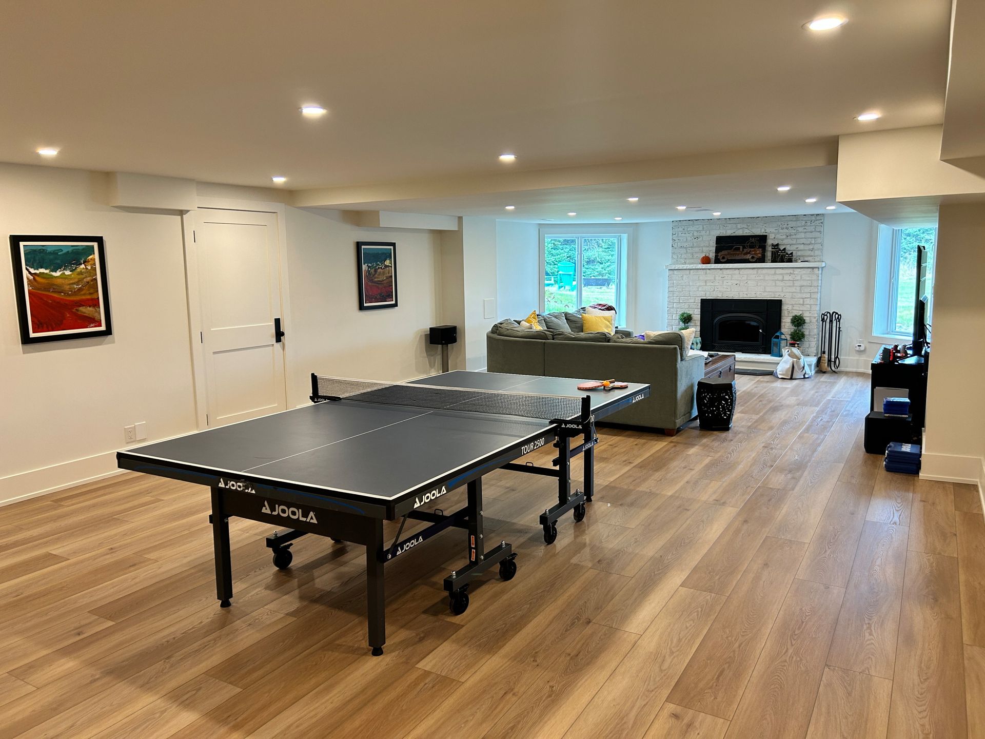 A finished basement with a ping pong table, sofa, fireplace, and hardwood floors.