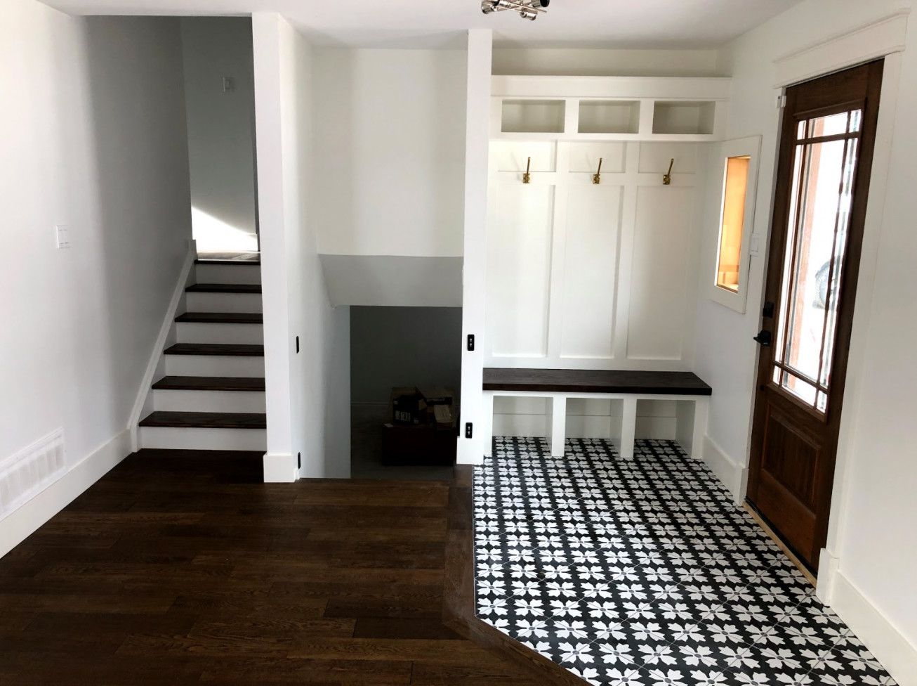 Entryway with dark wood floors, stairs, built-in bench with hooks, and patterned tile. White walls, brown door.