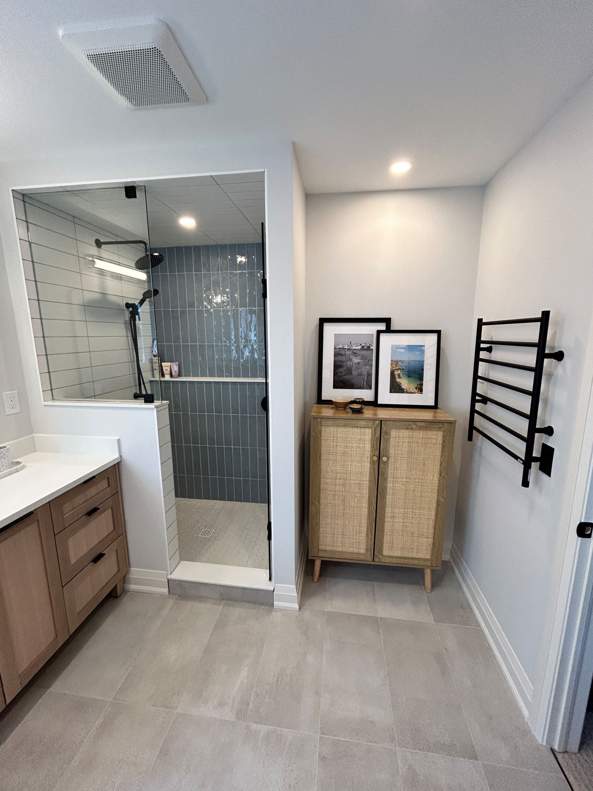 Bathroom with wood vanity, tiled shower, blue accent tile, woven cabinet, art, and black towel warmer.