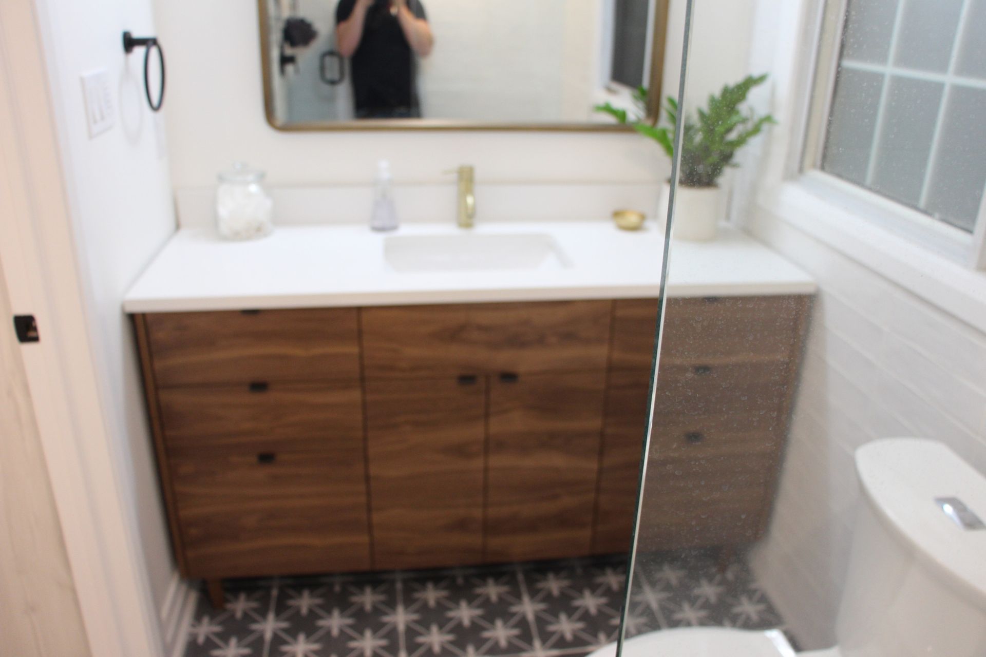 Modern bathroom with wooden vanity, white countertop, gold fixtures, and patterned floor tiles.