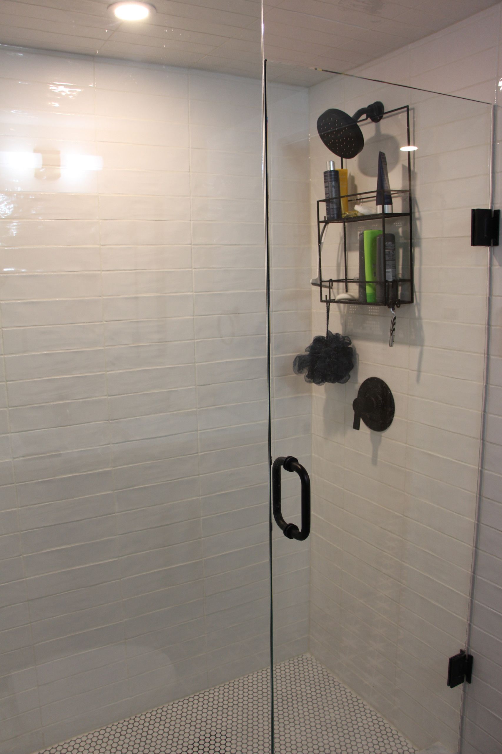 Modern shower with glass door and black fixtures. White tiled walls, dark shower caddy holding toiletries.