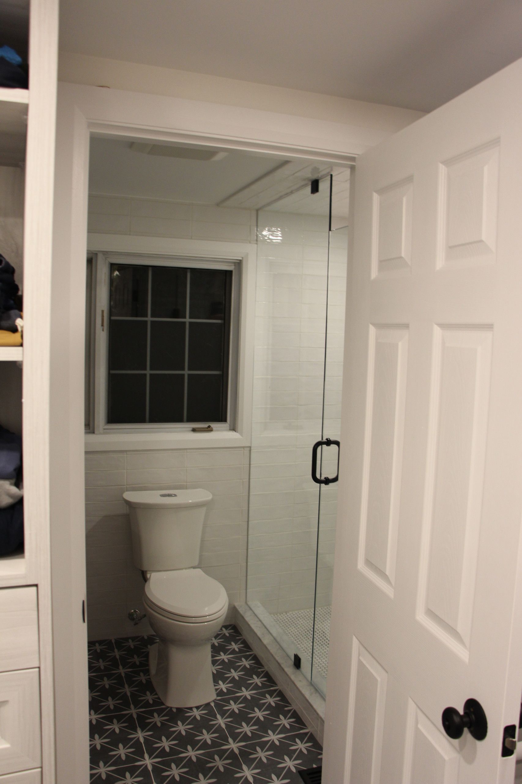 Small bathroom with toilet, glass shower, window, and patterned floor. White walls and door.