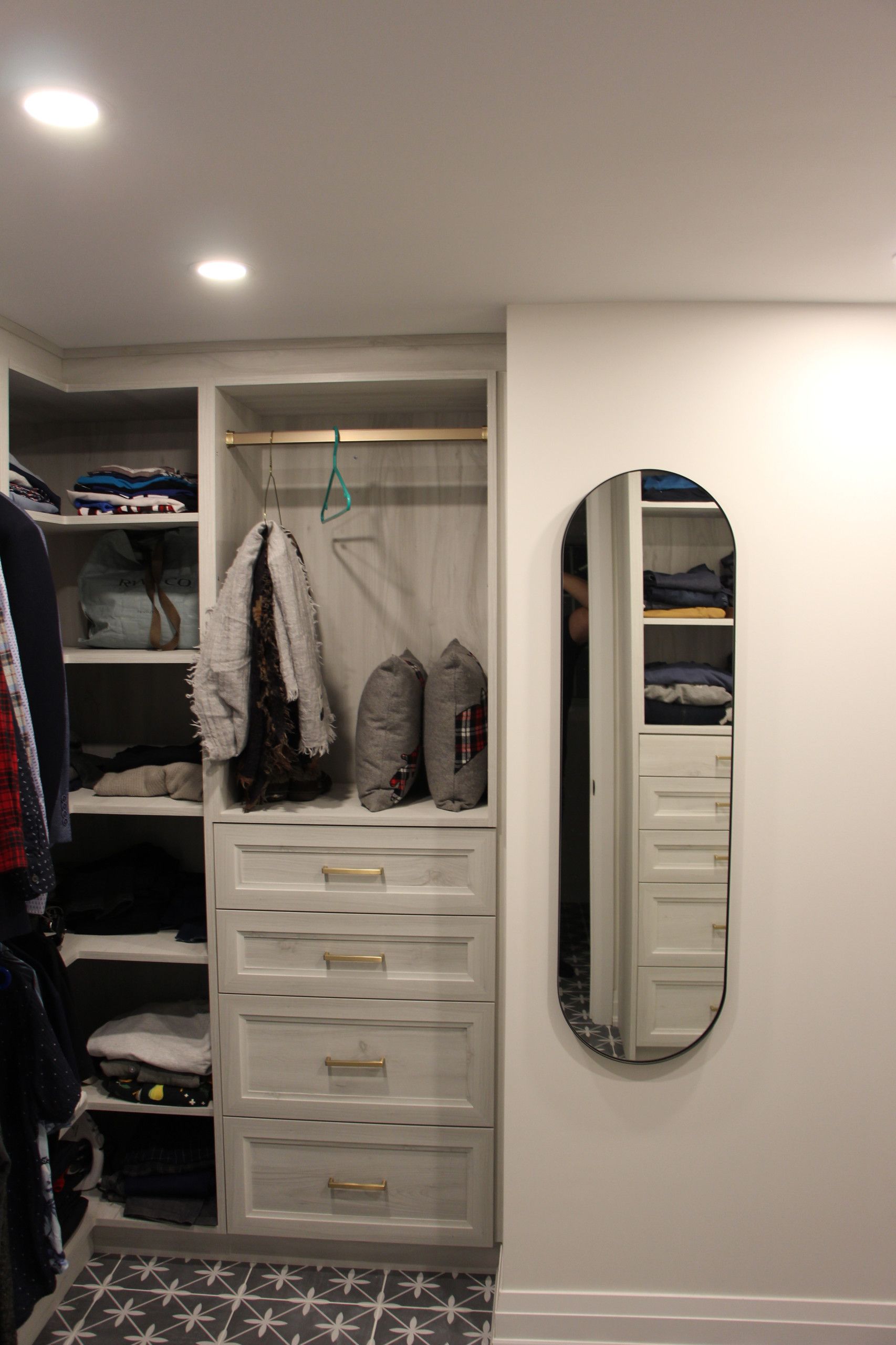 Walk-in closet with built-in shelves, drawers, and a mirror on a white wall; gray and gold accents.