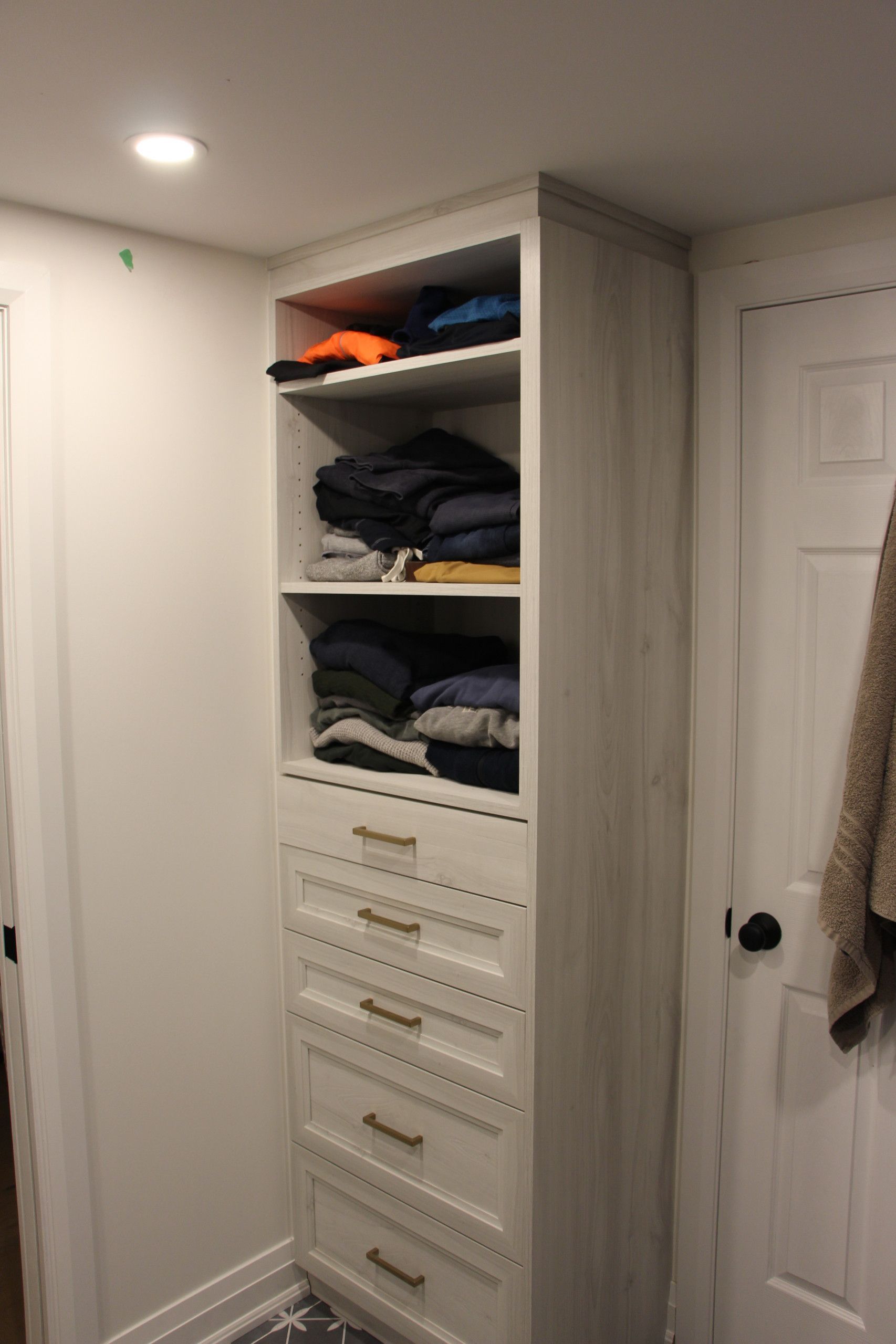 Tall, white closet unit with open shelving and drawers; holding folded clothing and located next to a door.