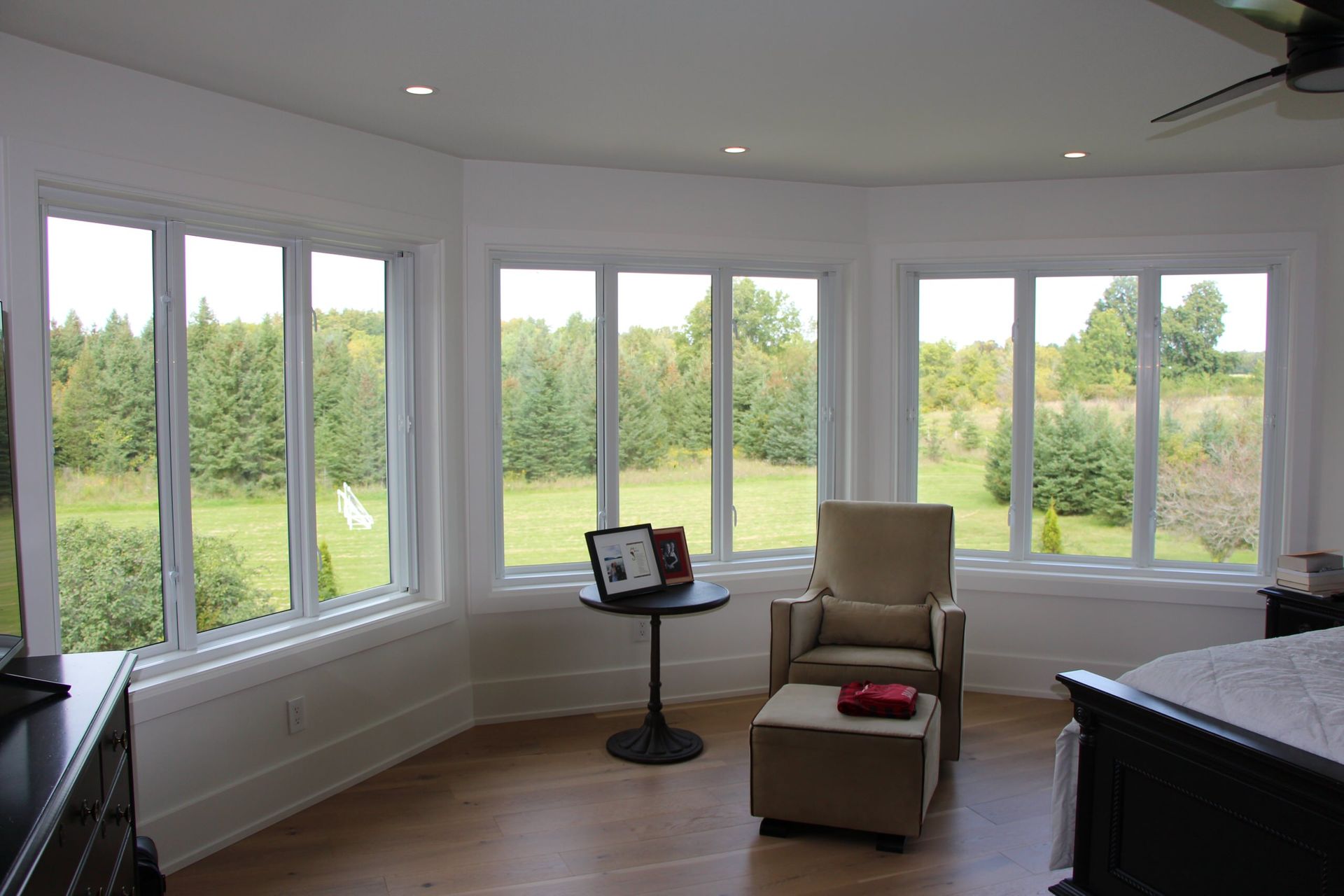 Bright bedroom with large windows overlooking a green field. A chair and side table are in front of the windows.