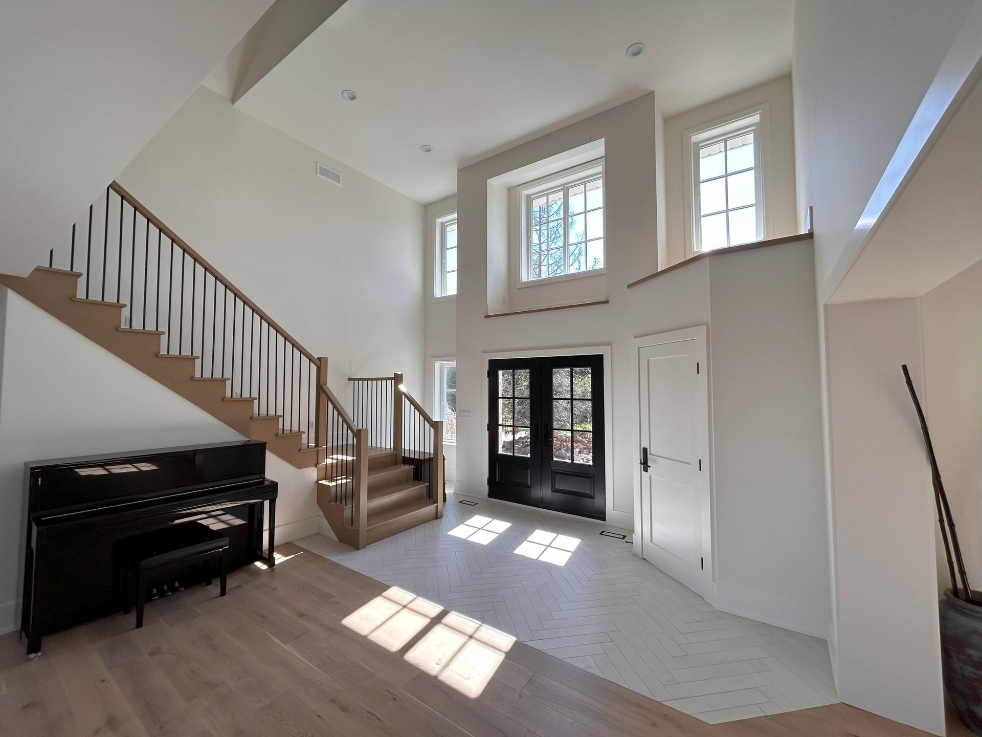 Spacious white foyer with staircase, black piano, and double black doors. Natural light streams in.