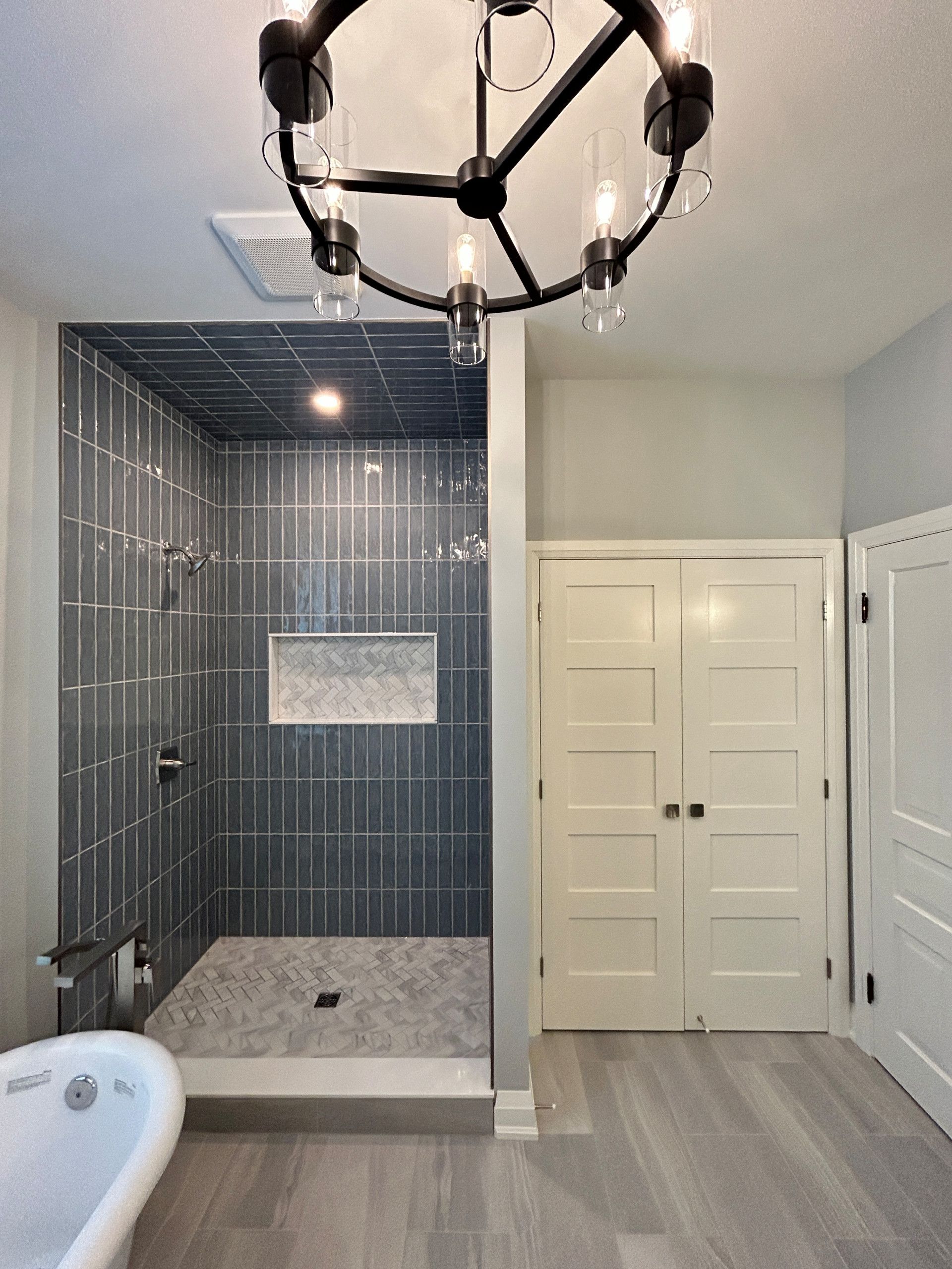 Modern bathroom with gray tiled shower, white door, chandelier, and gray wood-look floor.