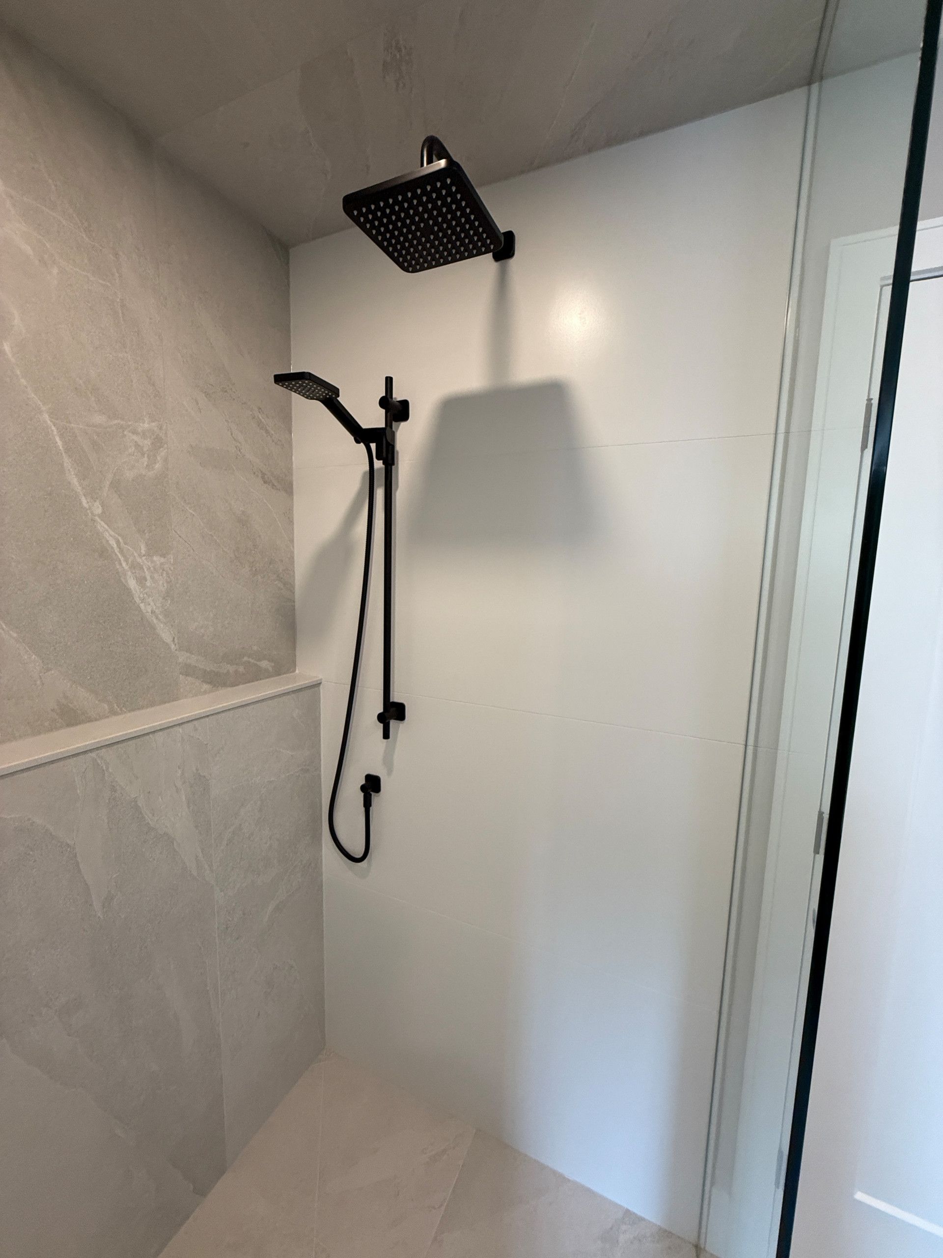 Black shower fixtures in a modern white-tiled shower with a stone accent wall and a glass door.