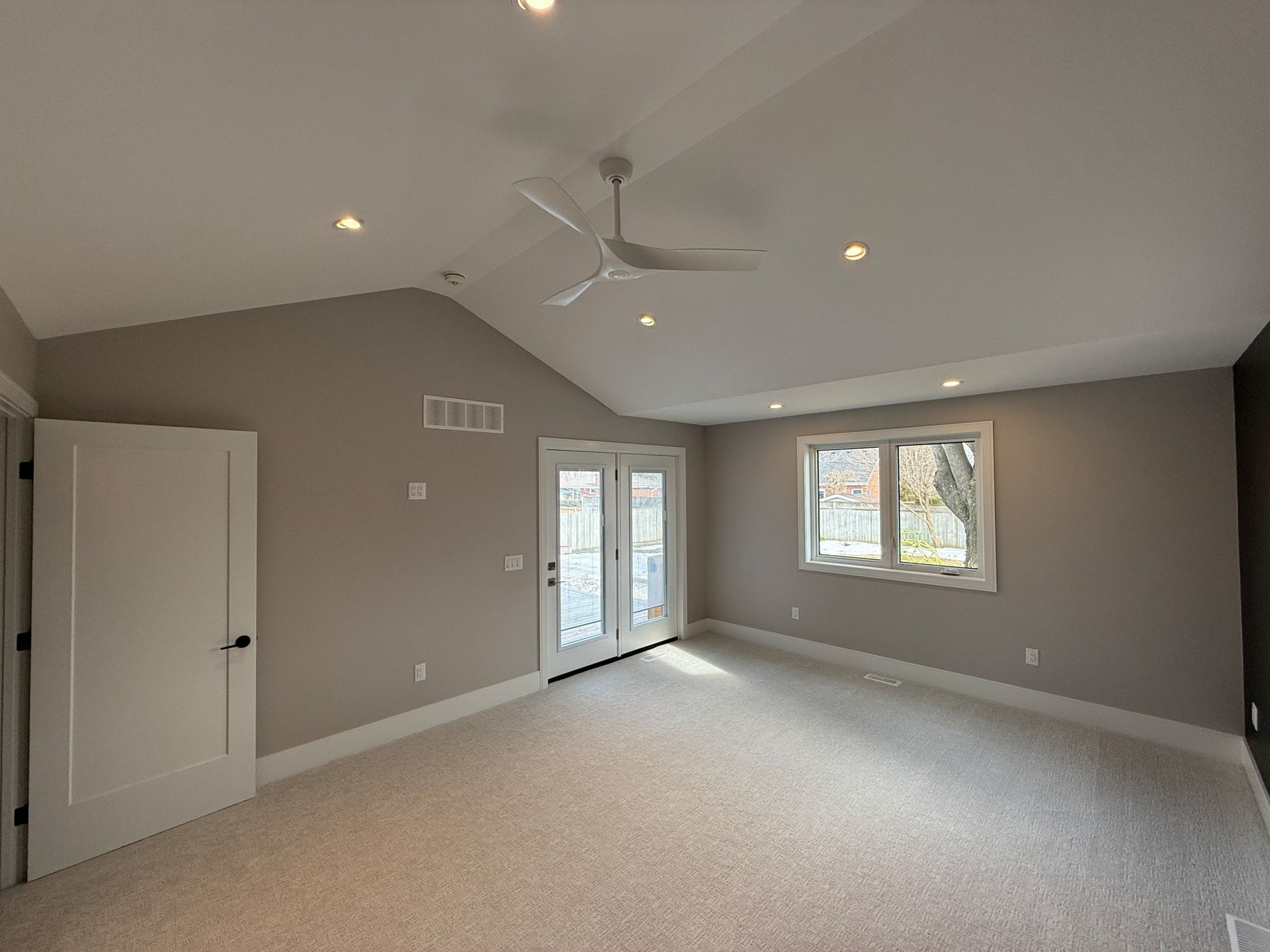 Spacious room with vaulted ceiling, ceiling fan, carpet, doors, and window. Walls are painted grey.