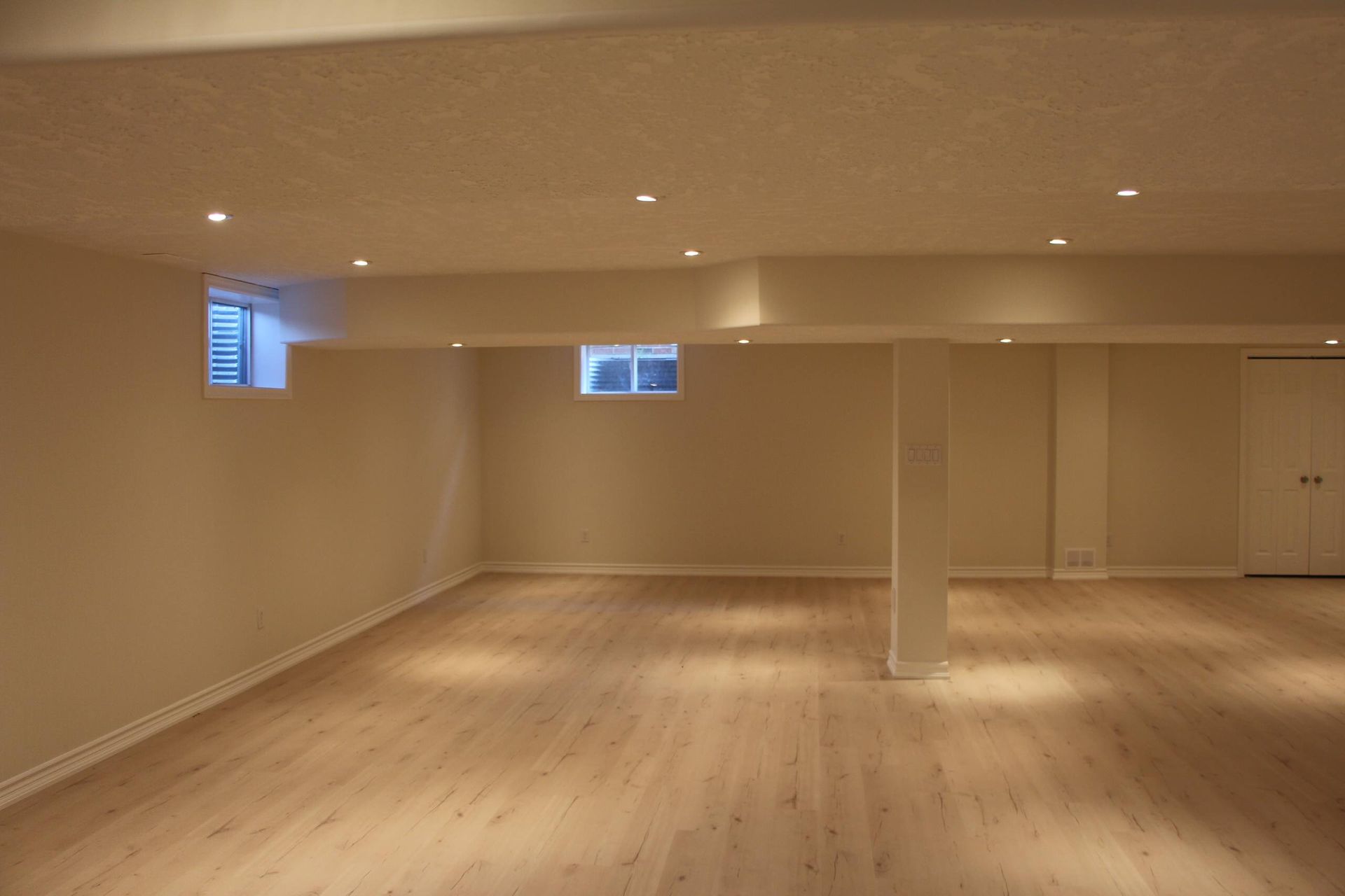 Empty basement room with beige walls, light wood floor, recessed lighting, and small windows.