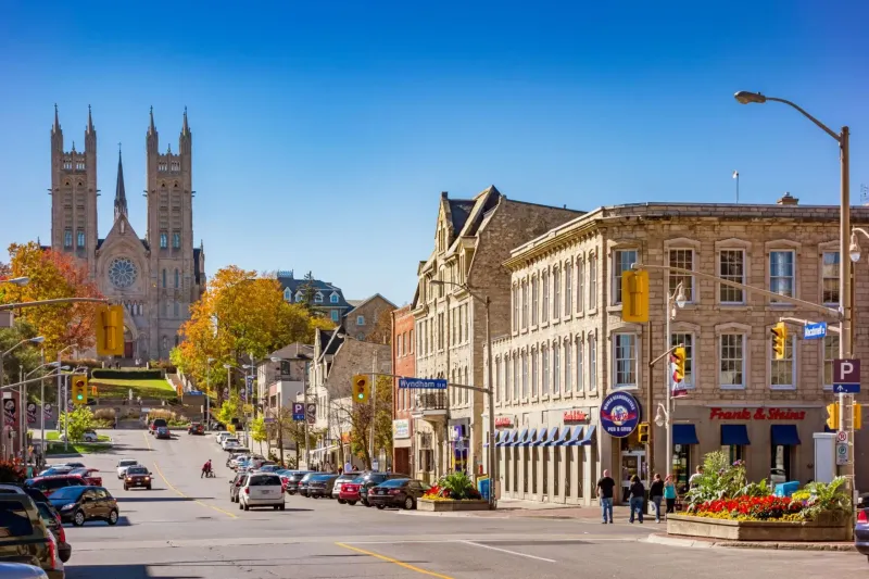 Street scene in bright sunlight with cathedral in the distance. Buildings line the street with cars, people and autumn trees.
