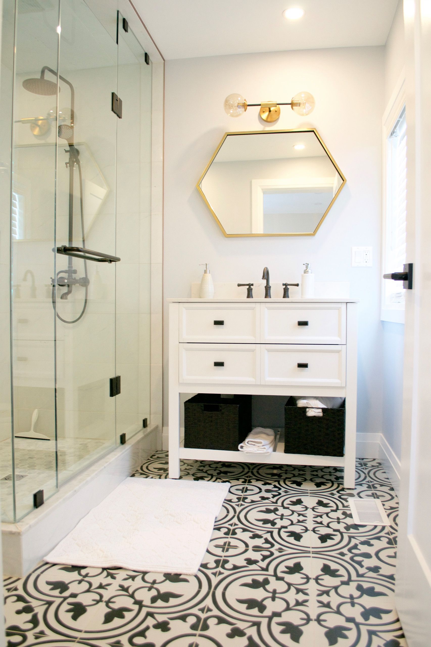 Bathroom with glass shower, patterned floor, white vanity, and gold-framed mirror.