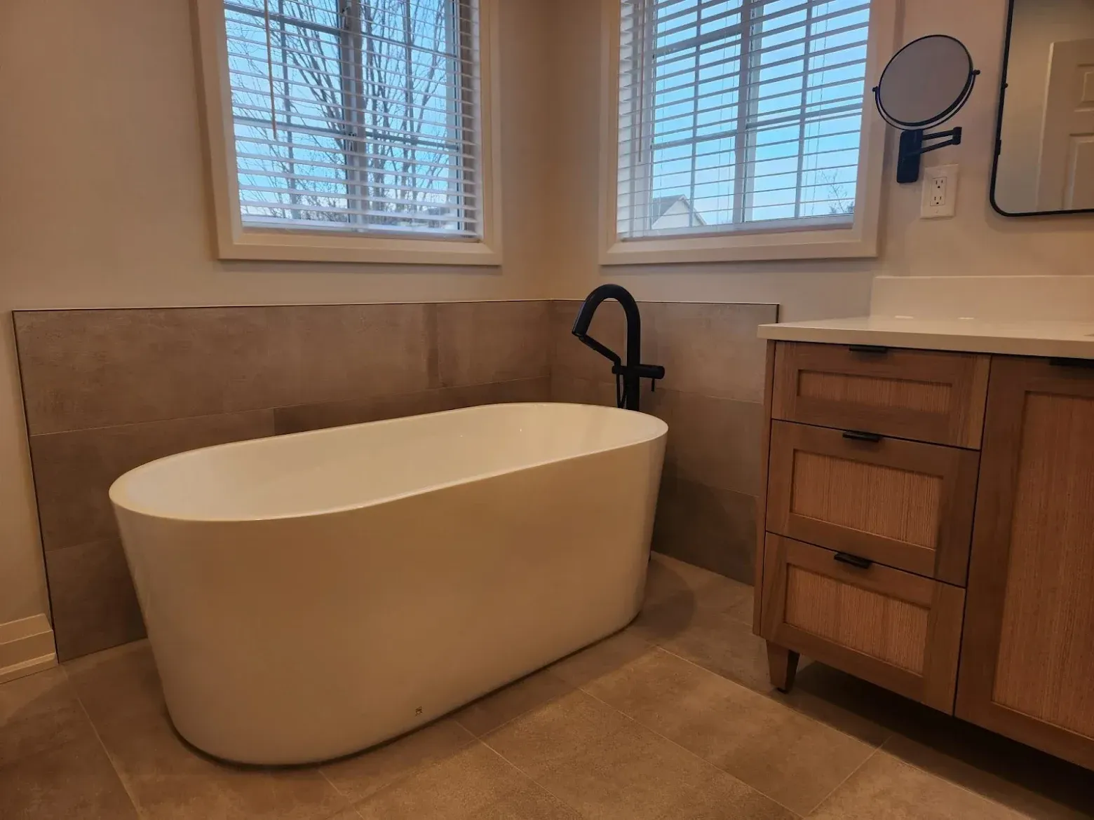 Freestanding white bathtub in a bathroom with a wooden vanity, windows, and neutral-toned tile.