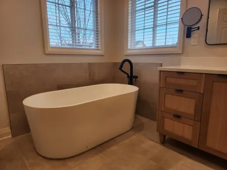 Freestanding white bathtub in a neutral-toned bathroom with two windows, and wood vanity.