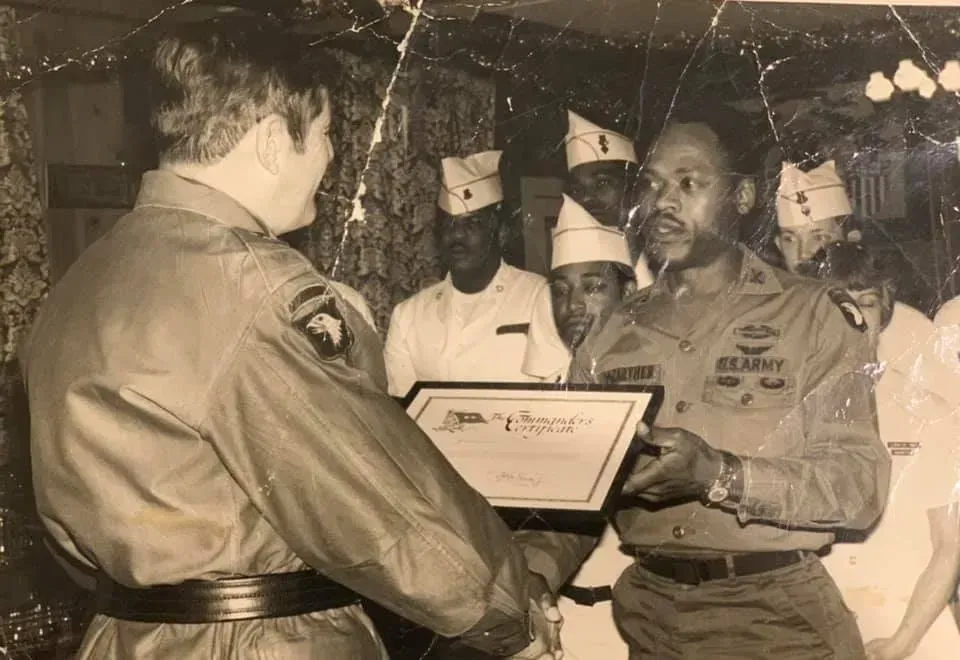 Everett Kelley with military personnel in uniform exchange a framed certificate during a formal indoor ceremony.