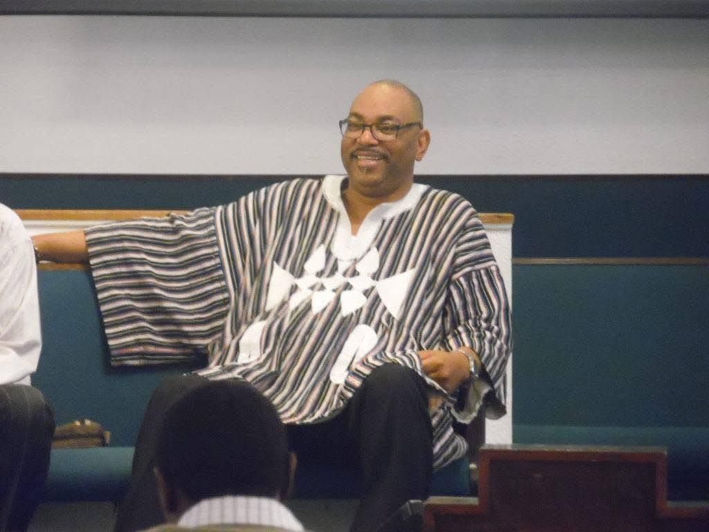 Everett Kelley in a striped dashiki sits on a bench in front of an audience, appearing to participate in a discussion.