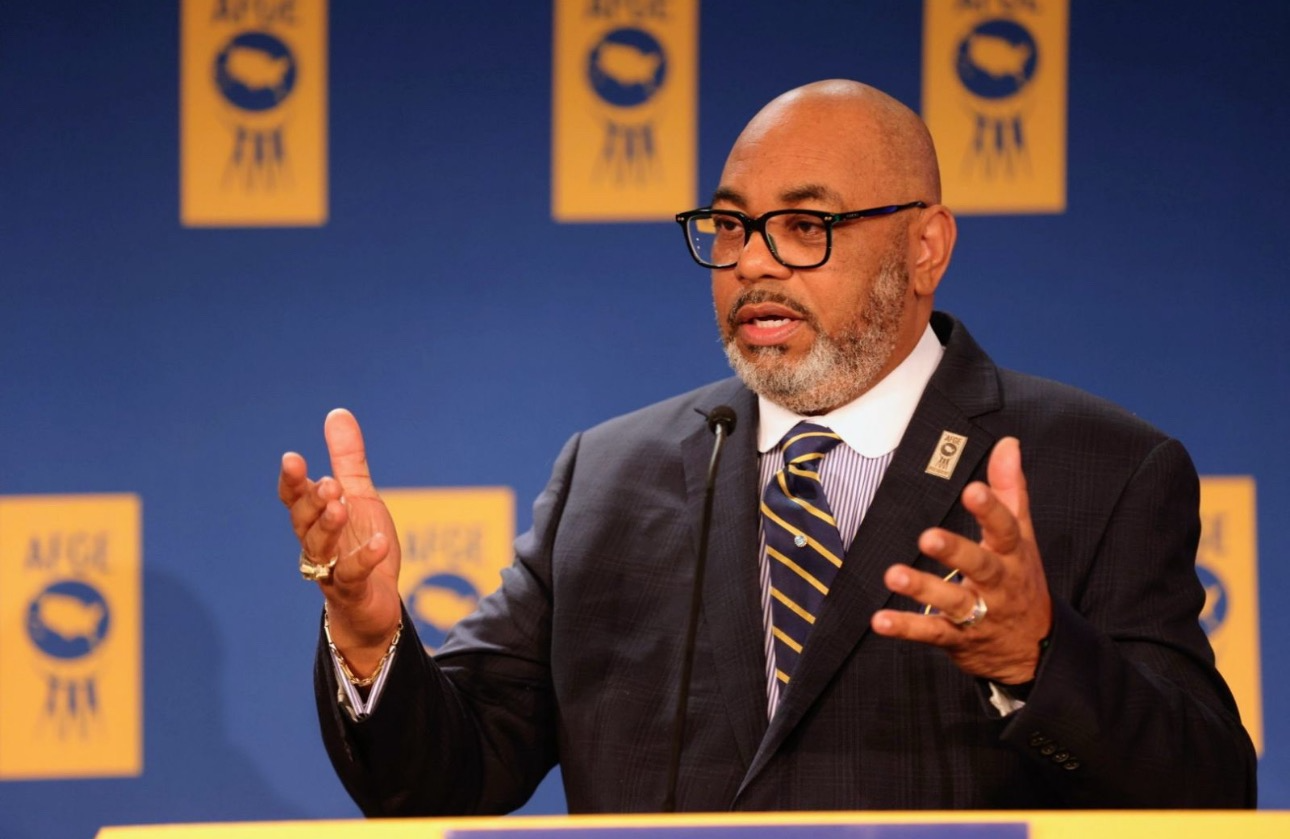 Everett B. Kelley in a suit and glasses gestures while standing at a podium in front of AFGE banners.