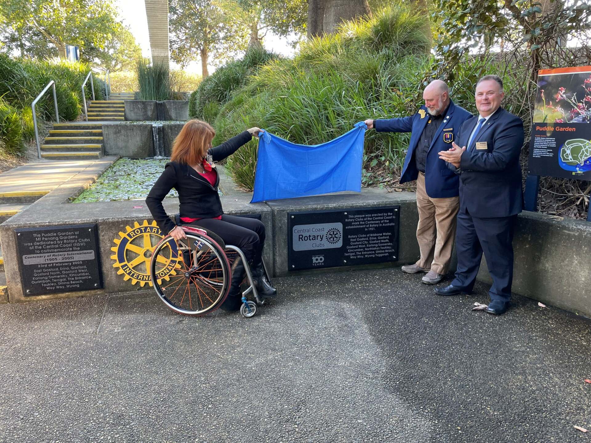 A woman in a wheelchair is holding a blue cloth while two men stand behind her.