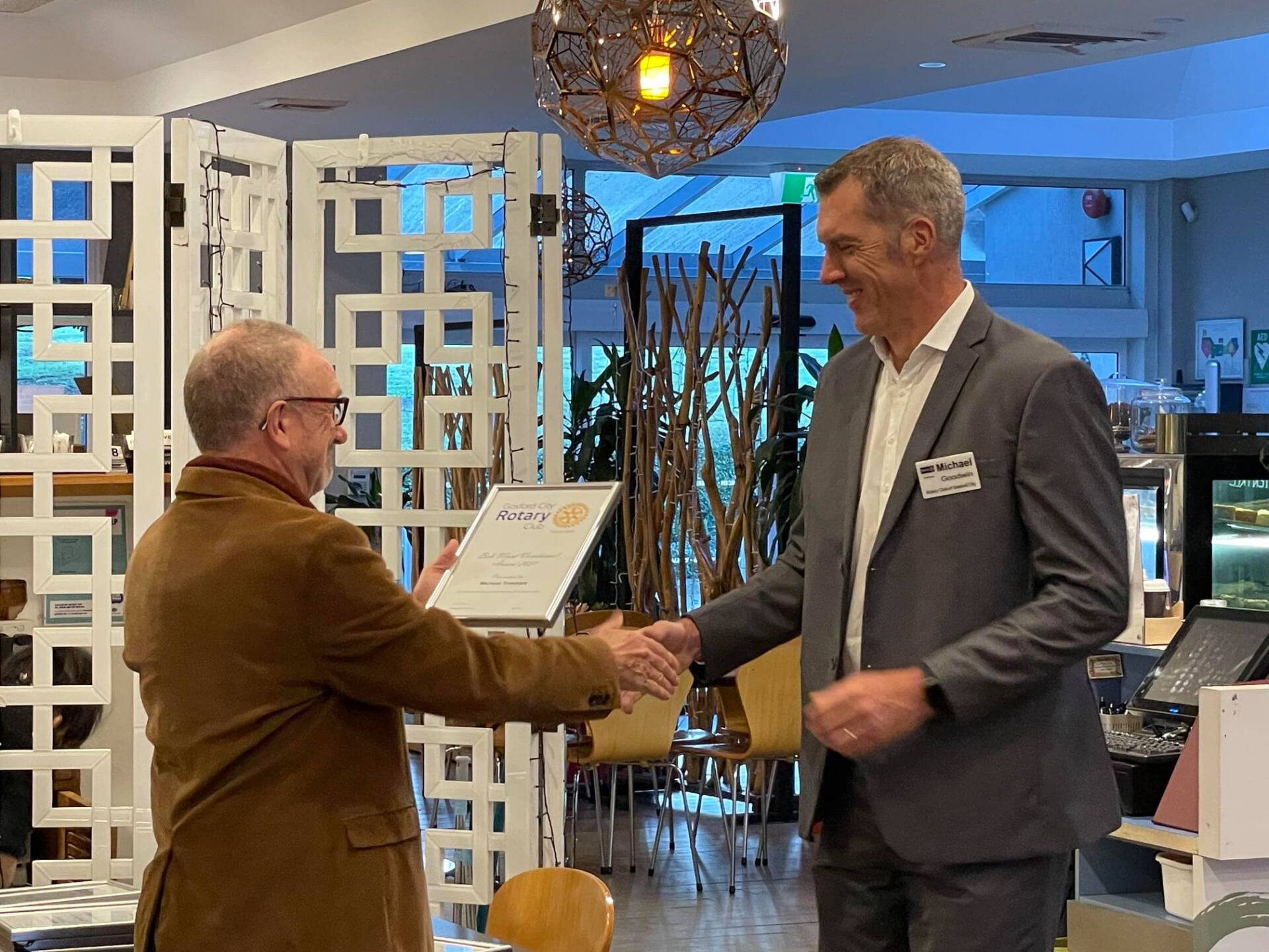 Two men are shaking hands in a restaurant while one man is holding a certificate.
