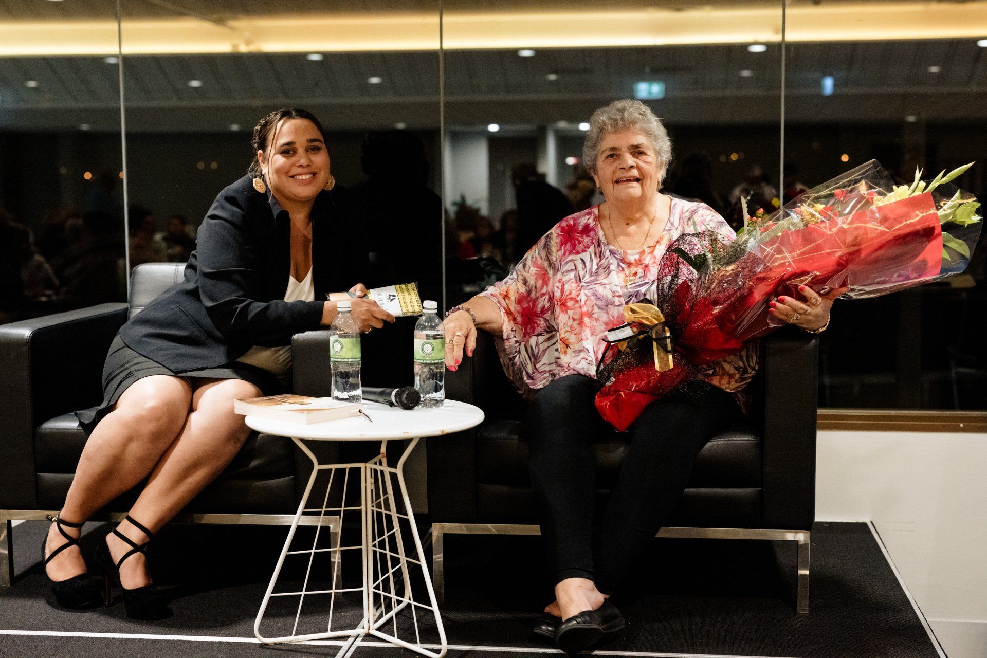 Two women are sitting on a couch and one is holding a bouquet of flowers.