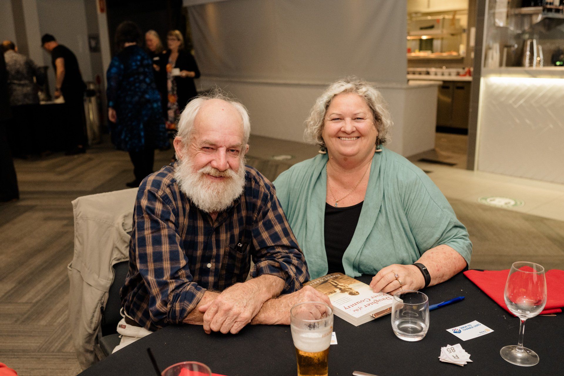 A man and a woman are sitting at a table with a book and a glass of beer.