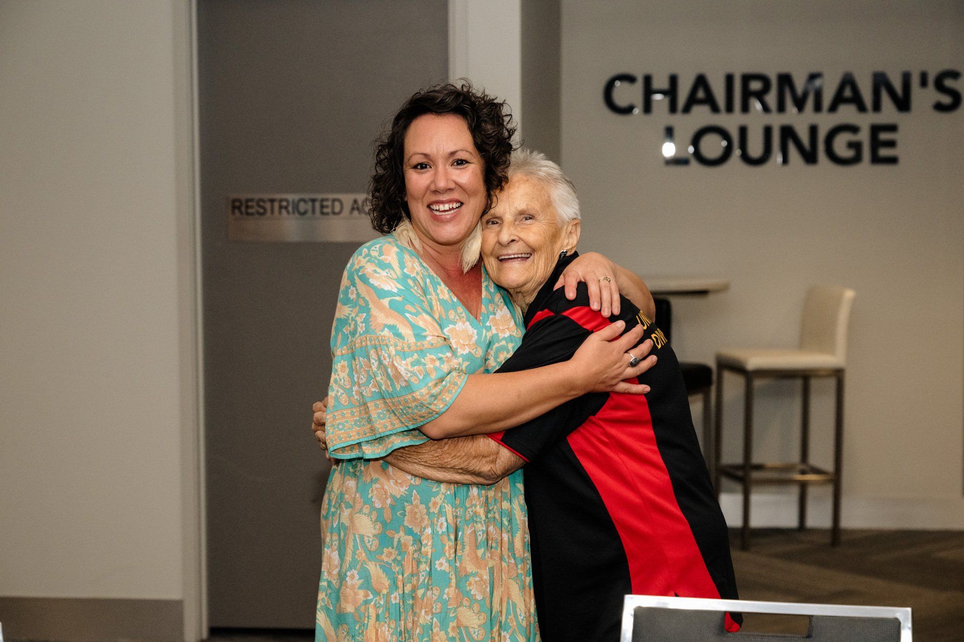 Two women are hugging each other in front of a sign that says chairman 's lounge.