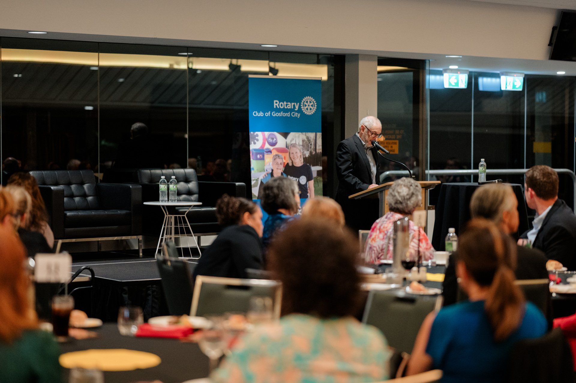 A man is standing at a podium giving a speech to a group of people sitting at tables.