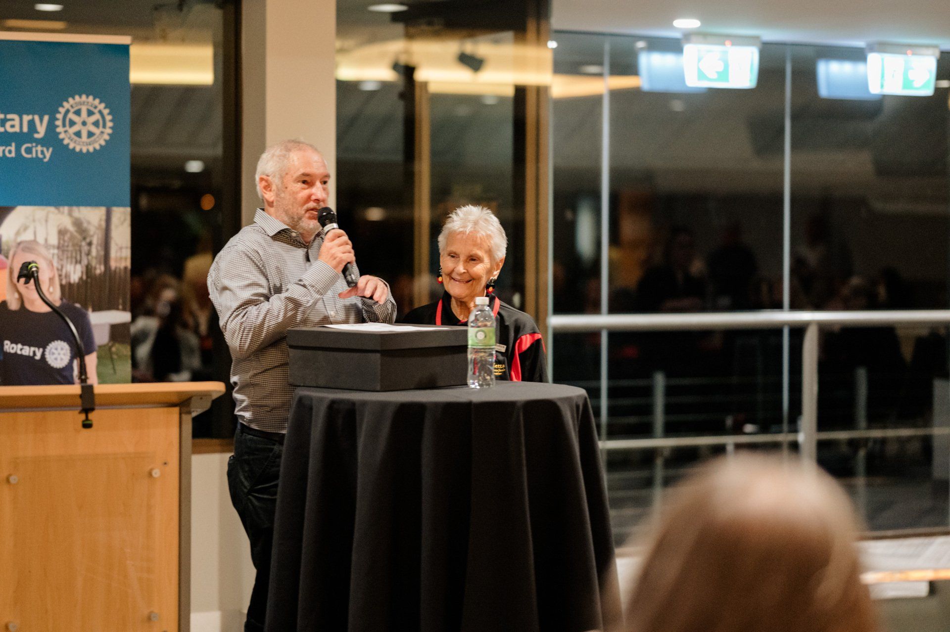 A man is standing at a podium talking into a microphone while a woman looks on.