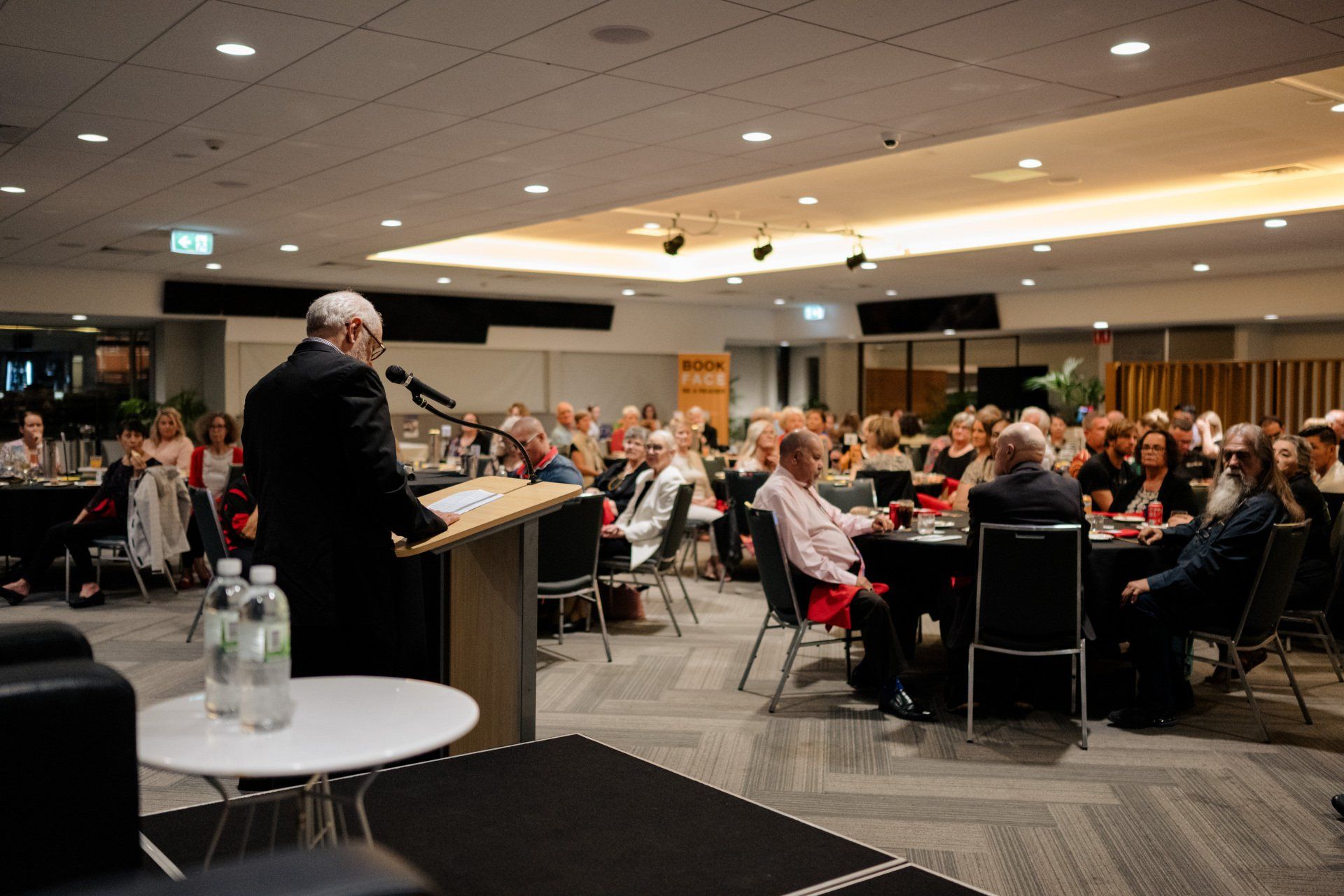 A man is standing at a podium giving a speech to a large group of people.