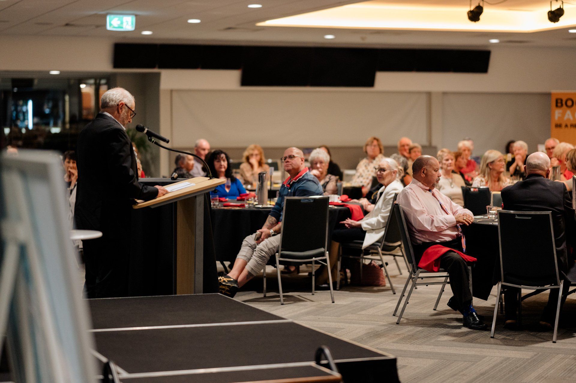 A man is standing at a podium giving a speech to a group of people sitting at tables.