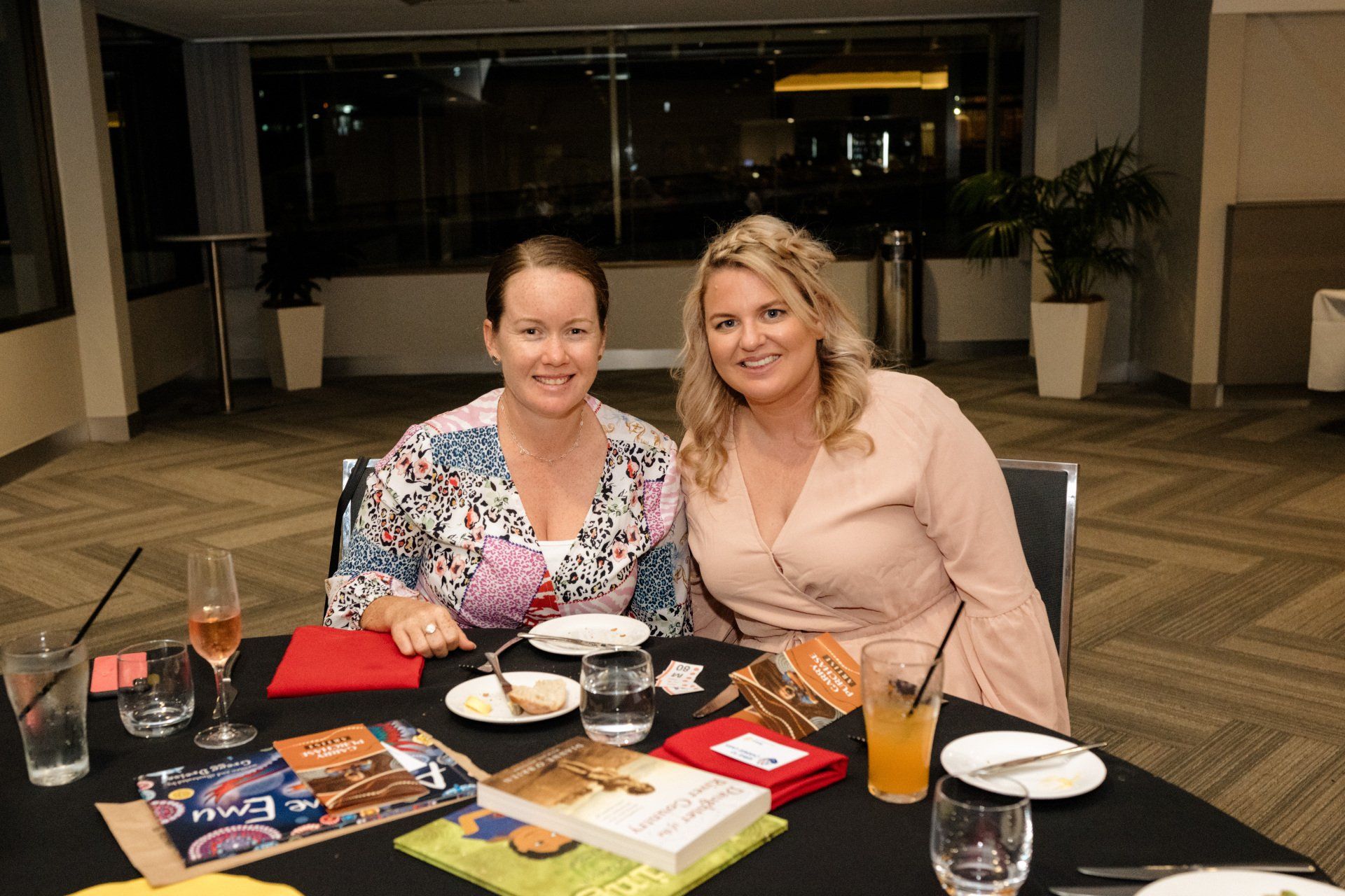 Two women are sitting at a table with drinks and books.