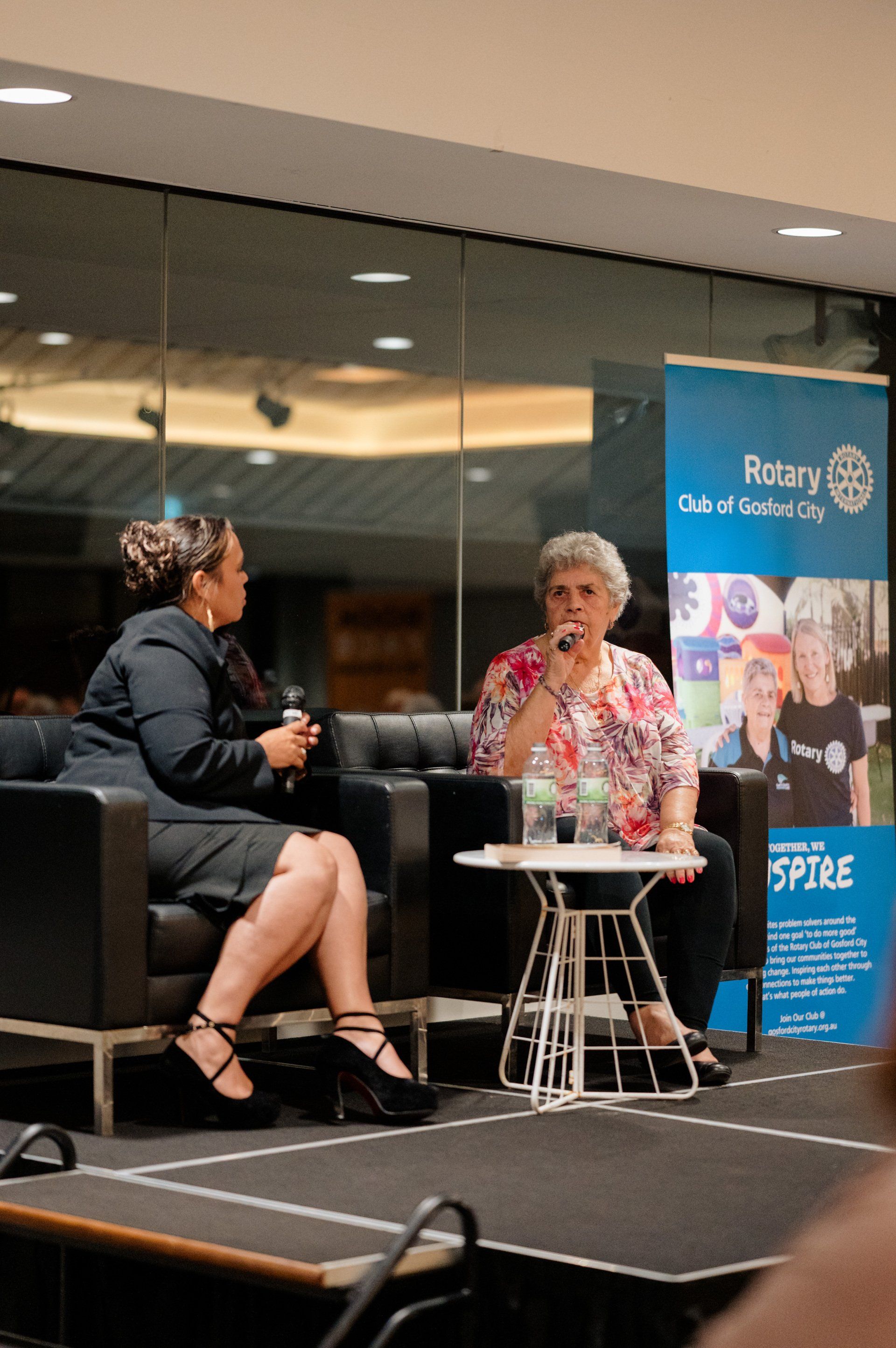 Two women are sitting in chairs on a stage talking to each other.