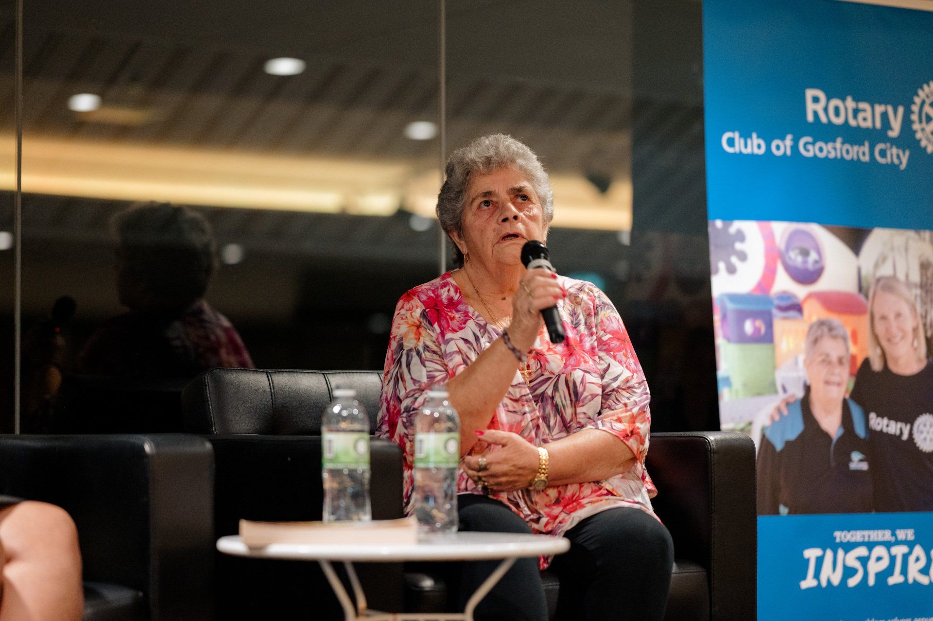A woman is sitting at a table talking into a microphone.