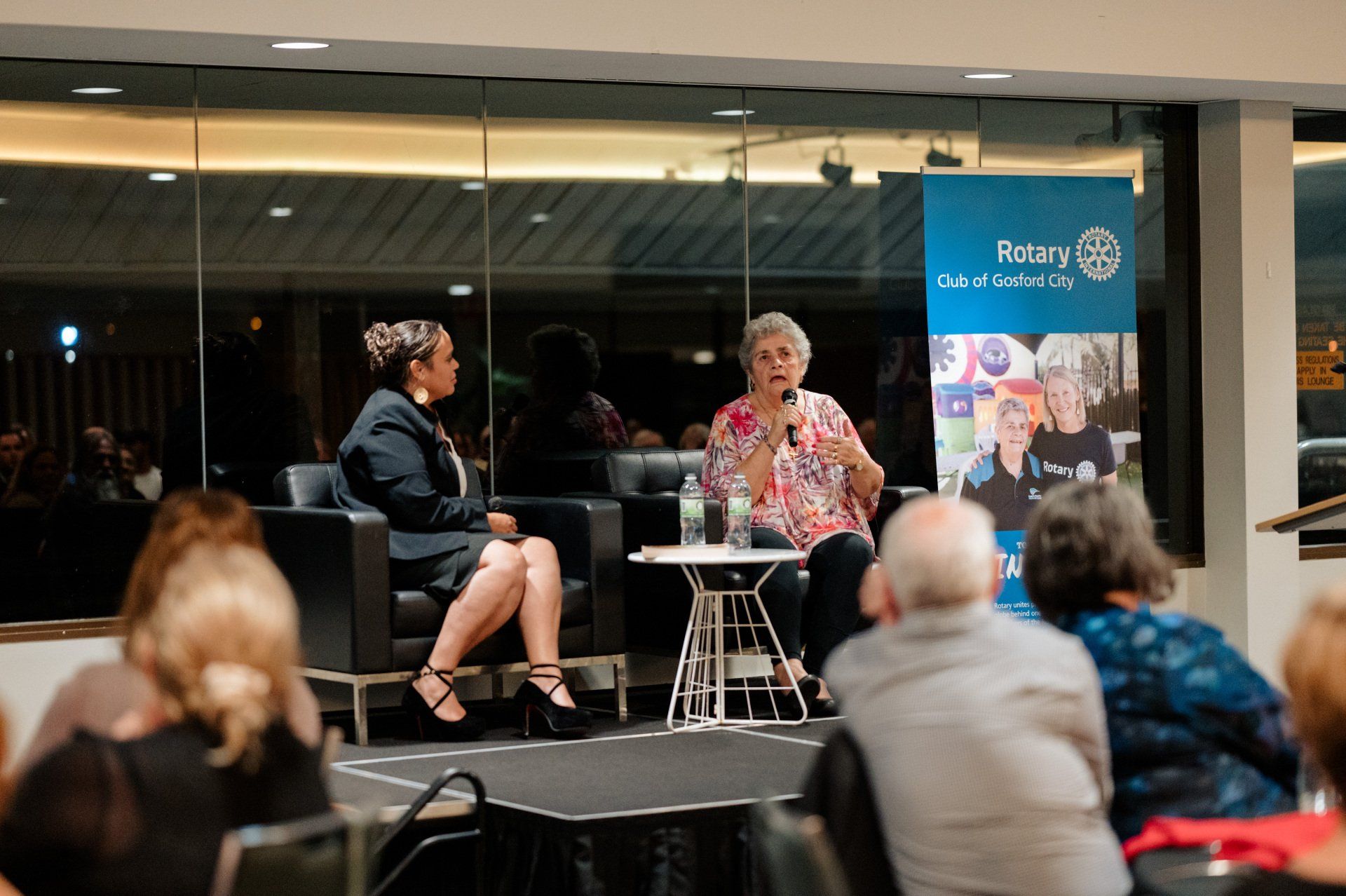 Two women are sitting at a table talking to a group of people.