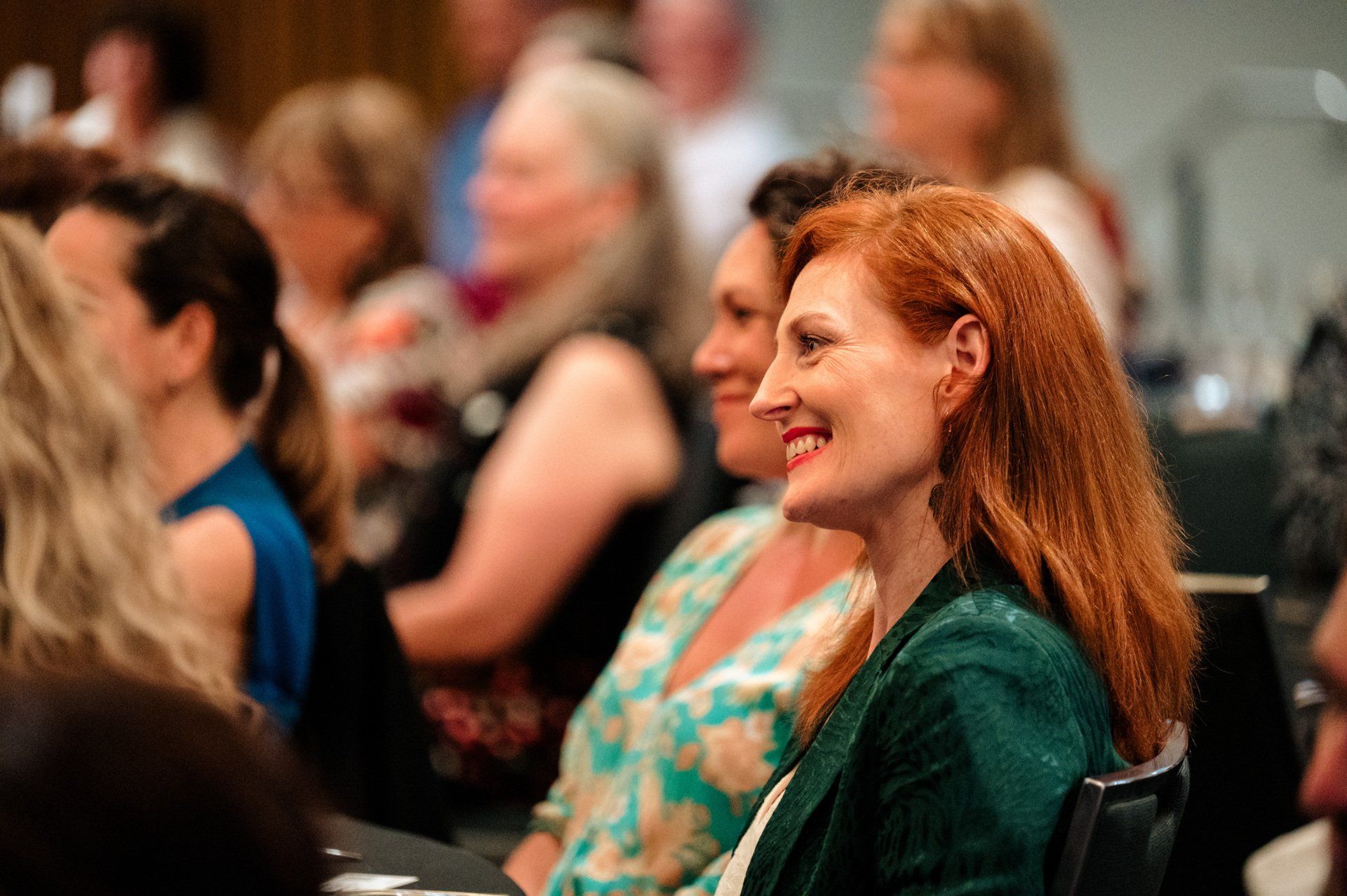 A woman with red hair is smiling while sitting in a crowd of people.