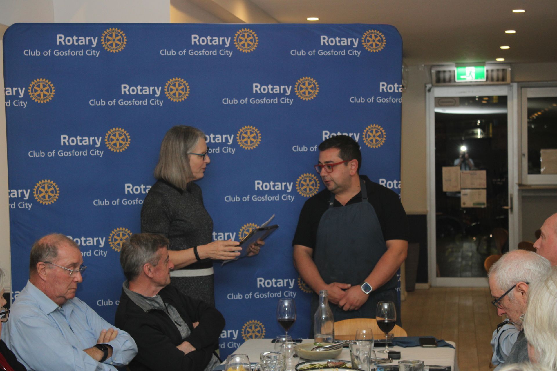 A group of people are sitting at a table in front of a wall that says rotary