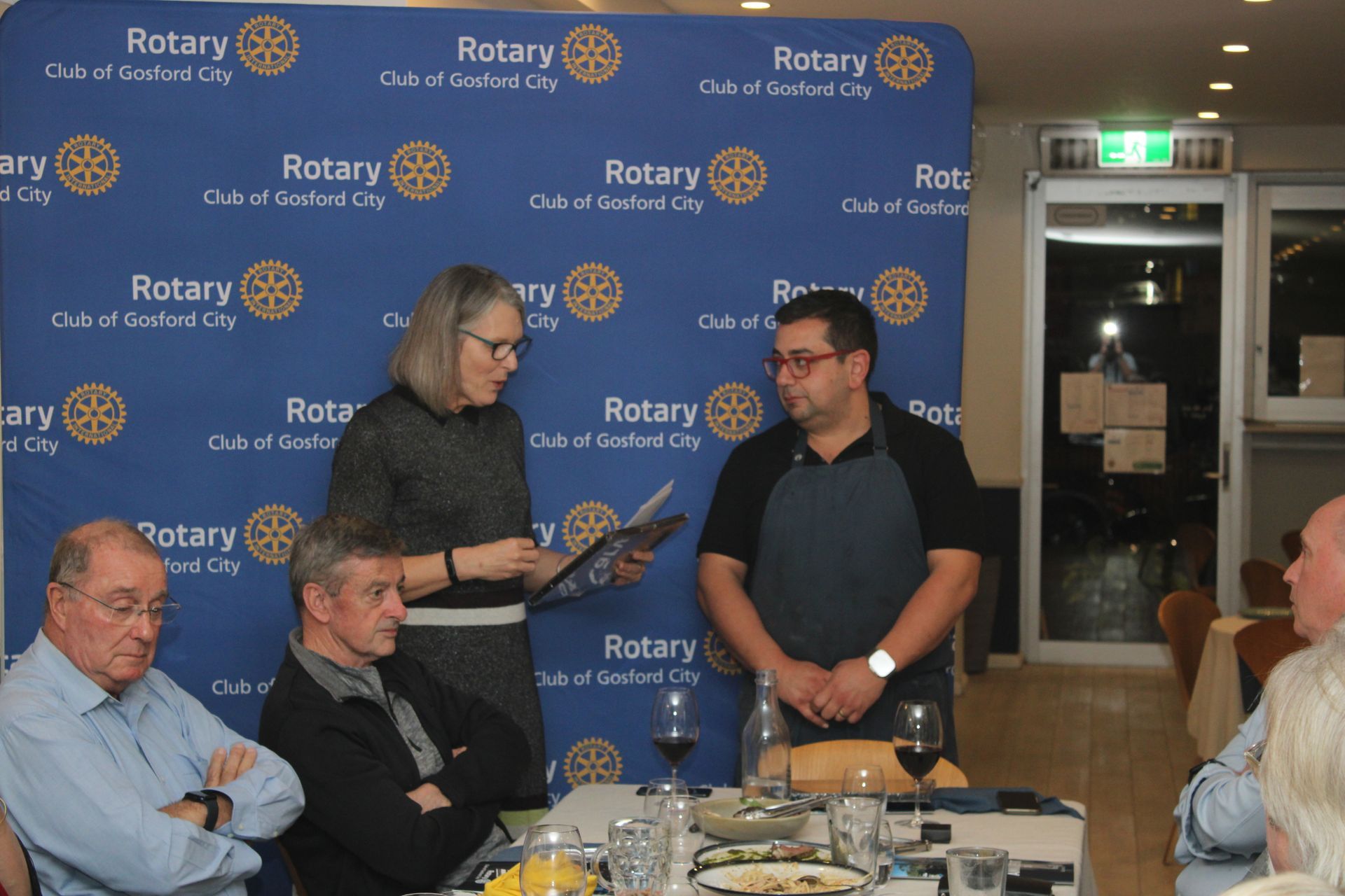 A group of people sitting around a table with a rotary sign behind them