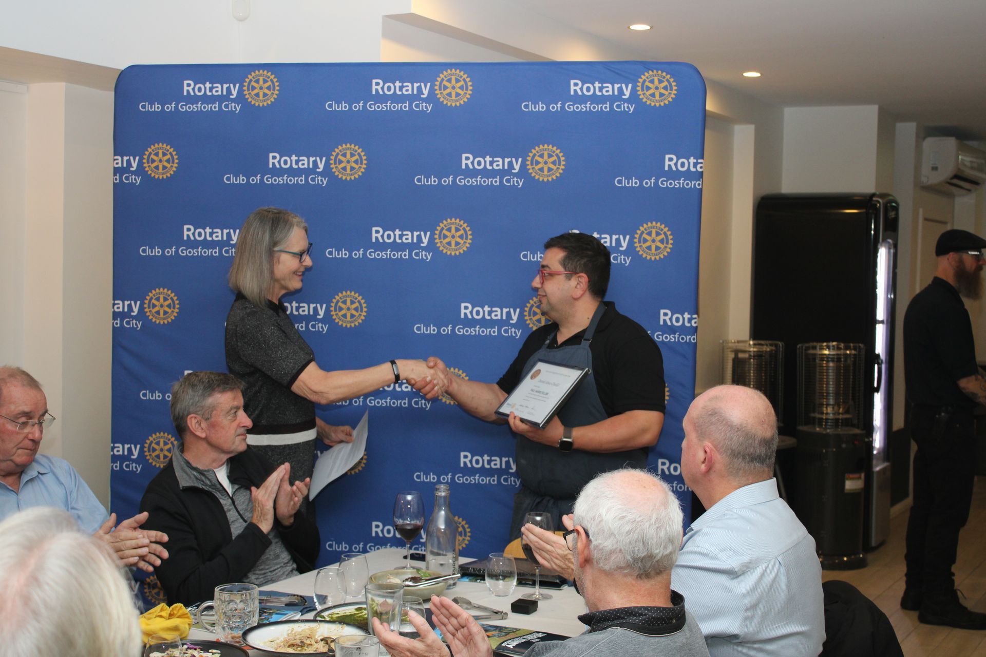 A group of people are sitting at a table and applauding while a man hands a woman a certificate.
